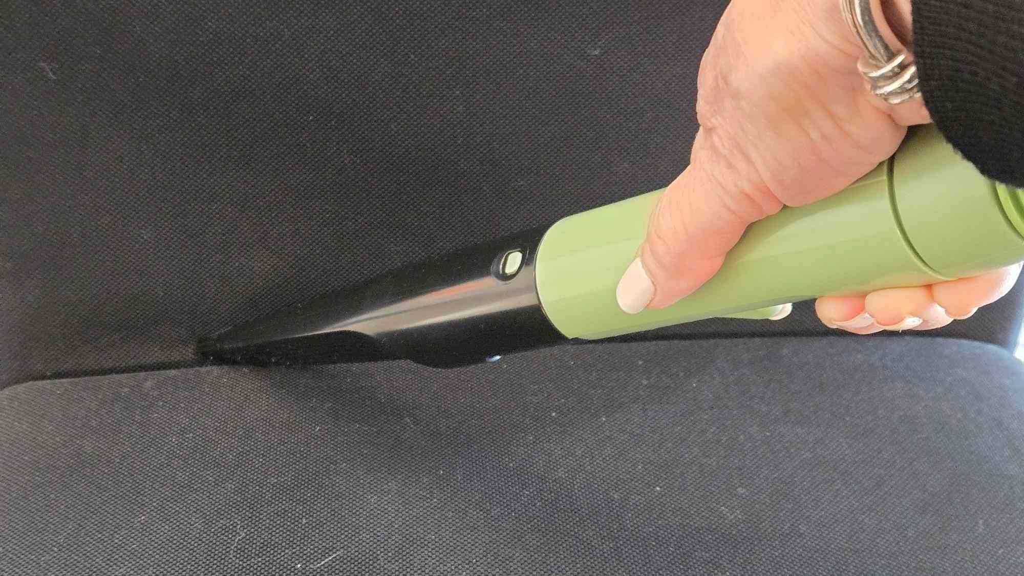 A hand guiding a narrow green cleaning nozzle into a dark car-seat seam in the source automotive-cleaner testing.