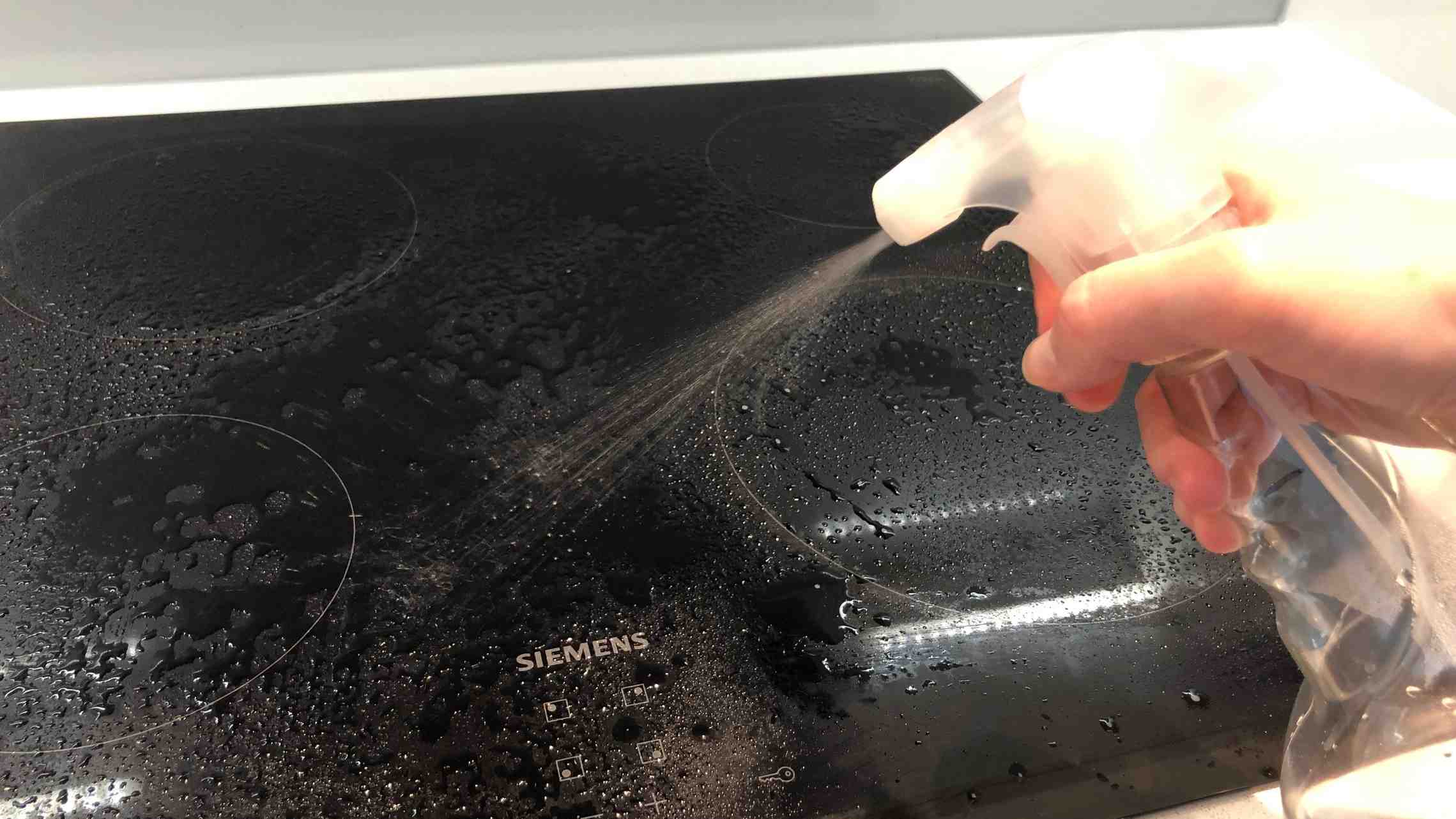 A hand spraying liquid over a black glass cooktop in the source cleaning demonstration; suitability still depends on the appliance instructions.
