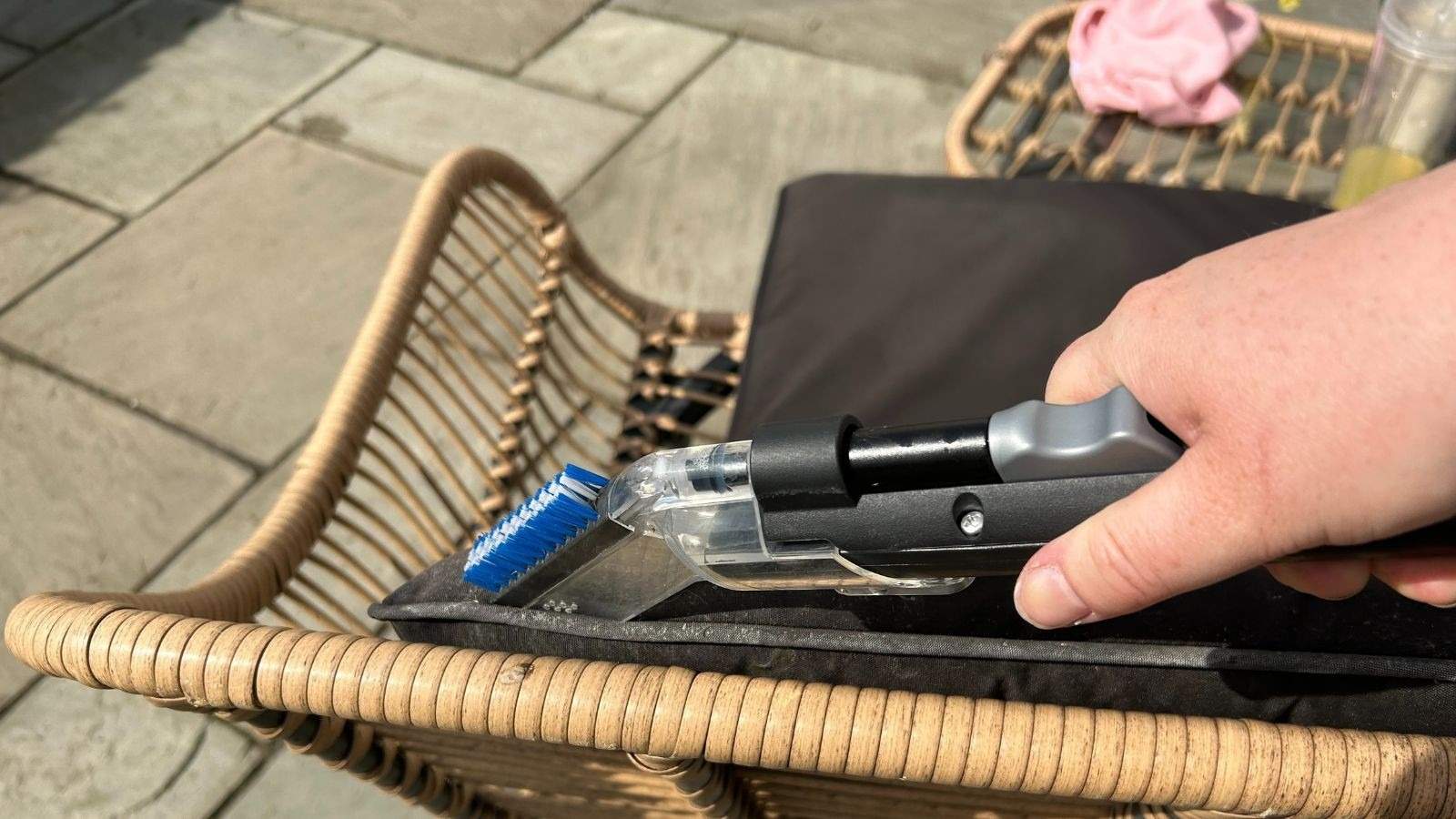 A hand guiding an upholstery-cleaning extraction head along a dark outdoor-seat cushion on a woven chair, as pictured in the source garden-furniture guide.