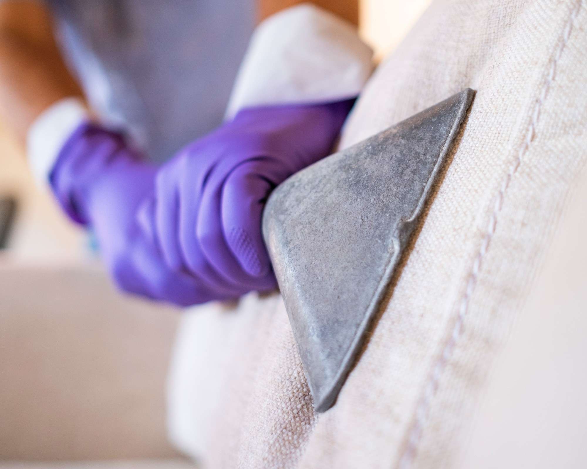 A gloved person guiding a transparent upholstery-cleaning head across a light fabric seat in the source guide; any machine treatment remains conditional on the furniture instructions.