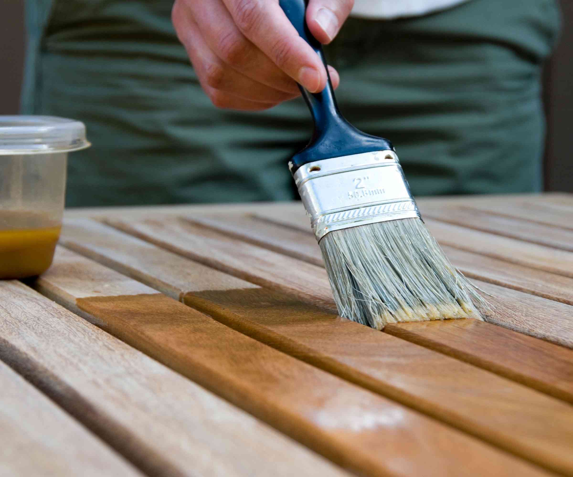A brush applying a wood treatment to a slatted outdoor table, illustrating the source discussion of color and protection.