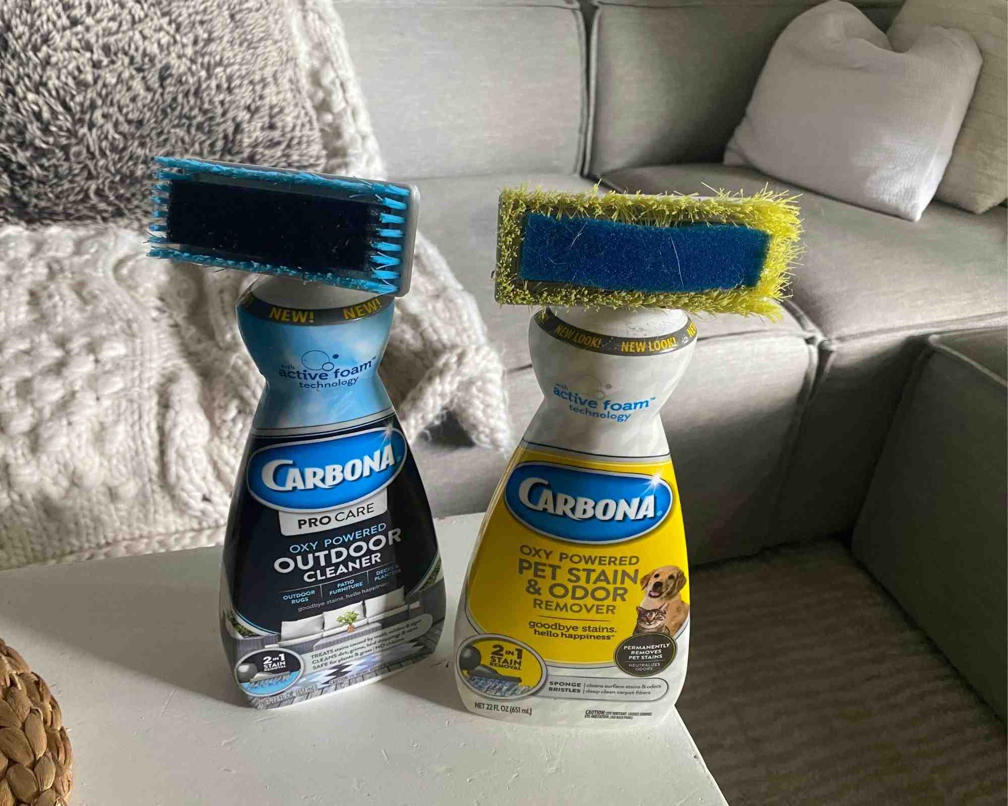 Two Carbona cleaning bottles with integrated brush heads on a table in front of a sofa, photographed for the source product comparison.